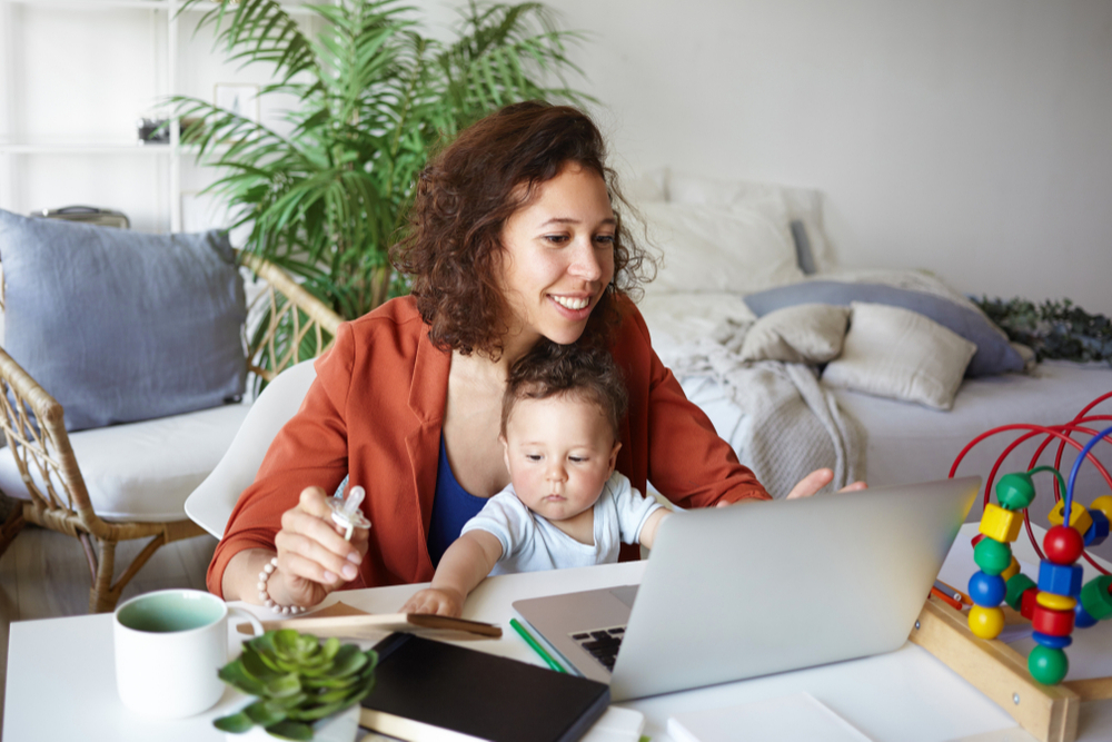 a mom and her baby on her lap in there living room at home with some toys around on the table. Mom is blogging on her computer. Large green plant in the background with coaches. 