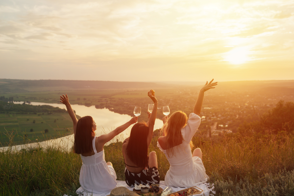 3 woman on a hillside with a river view and with wine glasses watching the sunset.